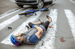 A man wearing a blue helmet lies injured on a pedestrian crossing next to his fallen bike, with a car nearby